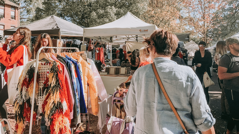 Bustling outdoor marketplace with white tents and diverse vendors displaying colorful clothing and attracting a crowd of shoppers on a sunny day