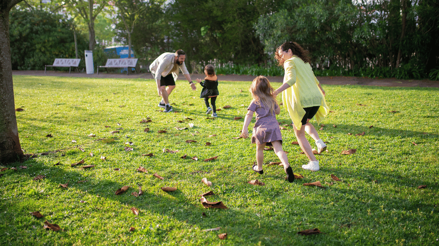 A group of people, appearing to be a family, are playing and interacting on a sunny grassy field with scattered trees