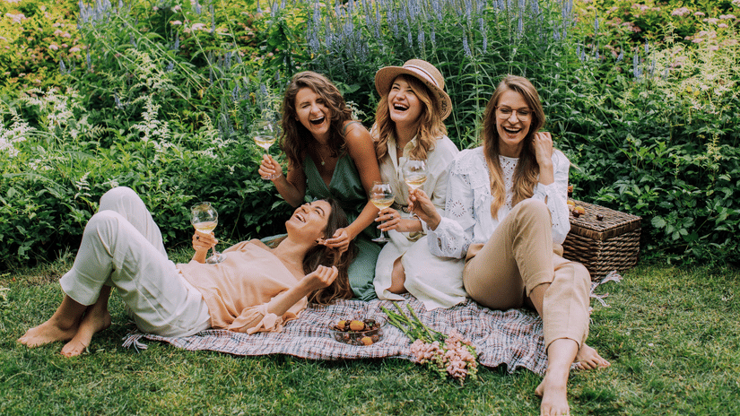 Four smiling women are enjoying a picnic on a blanket in a lush garden filled with colourful flowers and greenery