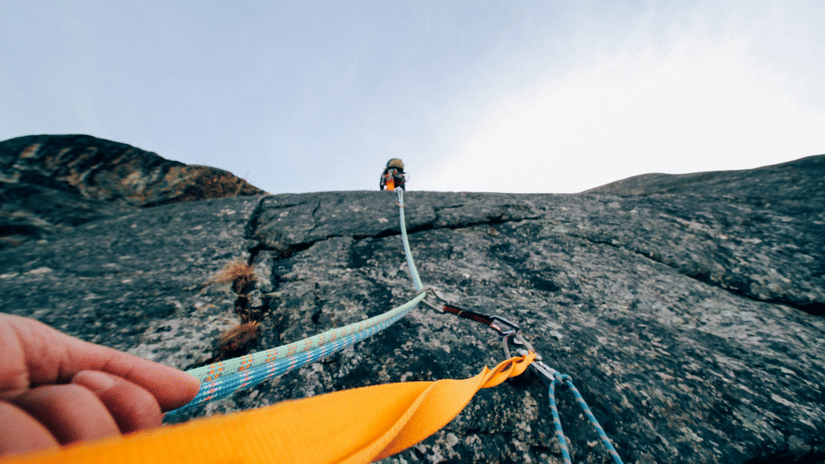 Rock climber ascending a steep cliff face, secured by ropes and with another climber visible below. A bright orange tarp is visible
