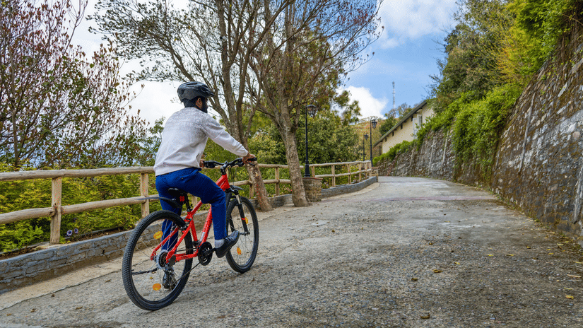 A cyclist enjoys a leisurely ride on a winding paved path, surrounded by lush greenery and trees under a partly cloudy sky - Digantaa Resort in Mukteshwar