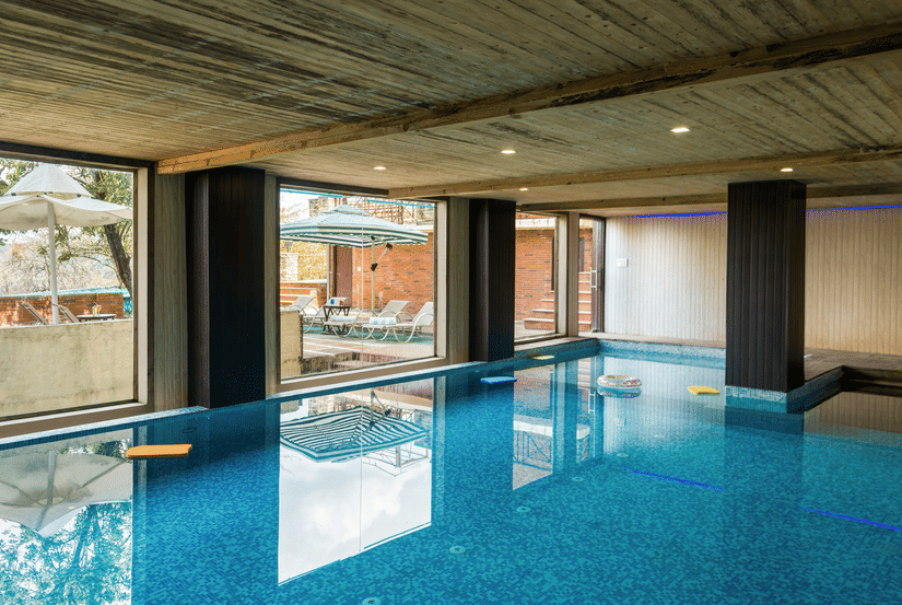An indoor swimming pool with clear blue water, surrounded by large windows looking out onto greenery