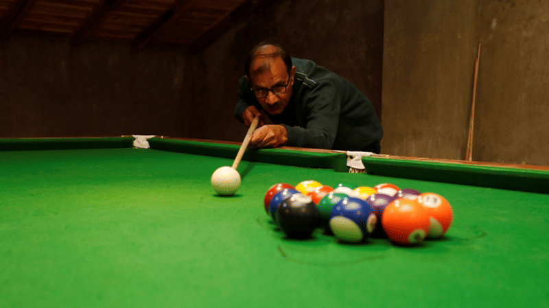 A person playing billiards, aiming a cue stick at colourful balls on a green pool table.