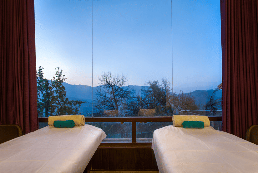Two massage beds in a spa room with large windows at Digantaa Resort in Mukteshwar providing a tranquil view of the mountains