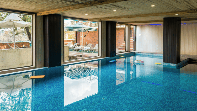 An indoor swimming pool with clear blue water, surrounded by large windows looking out onto greenery