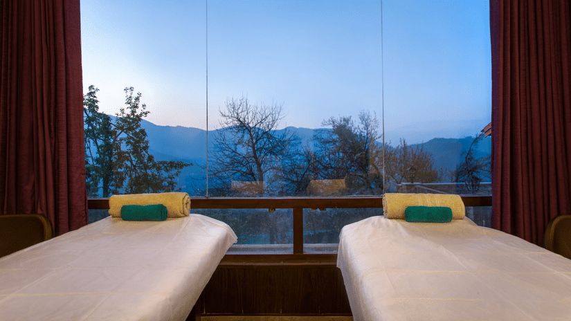Two massage beds in a spa room with large windows at Digantaa Resort in Mukteshwar providing a tranquil view of the mountains