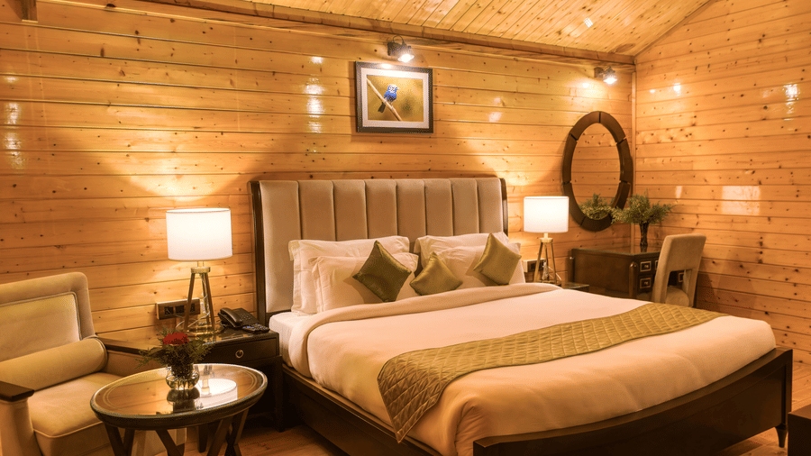 A cosy hotel room of Digantaa Resort in Mukteshwar featuring  wooden walls and ceiling. A large bed with a striped comforter, two pillows, bedside table with a lamp and a wooden armchair