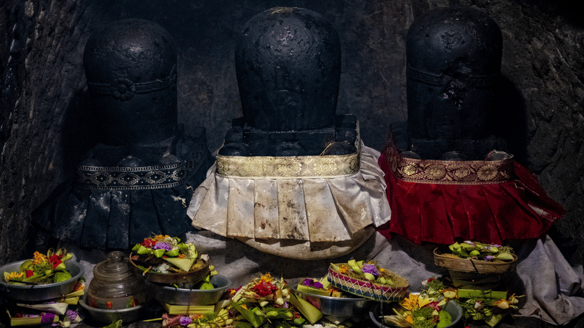 An ancient stone shrine with a dark, textured wall. Offerings of food and flowers are neatly arranged on a white cloth in front of a statue